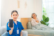 © CandyRetriever  - Female caregiver holding mobile phone, showing device screen with senior woman patient suffering from heart attack on bed background. Elderly people emergency health crisis need hospital medical help.