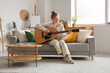 © Pixel-Shot - Young woman playing guitar on sofa in living room