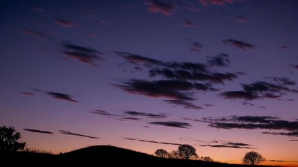 Wall Mural - Time lapse footage of a colorful twilight sky transitioning into morning or evening over hills and silhouetted trees with vivid cloud formations and atmospheric lighting perfect for seasonal nature