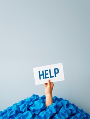 Poster - Hand holding HELP sign above pile of blue crumpled paper on light background expressing stress or overload in creative business concept.
