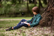 © Cavan - Child boy reading a book in the park leaning against an old tree. N