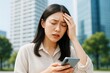 © Pixels Hunter - Stressed woman holding smartphone outdoors with eyes closed and hand on forehead, showing frustration or worry in urban background setting.