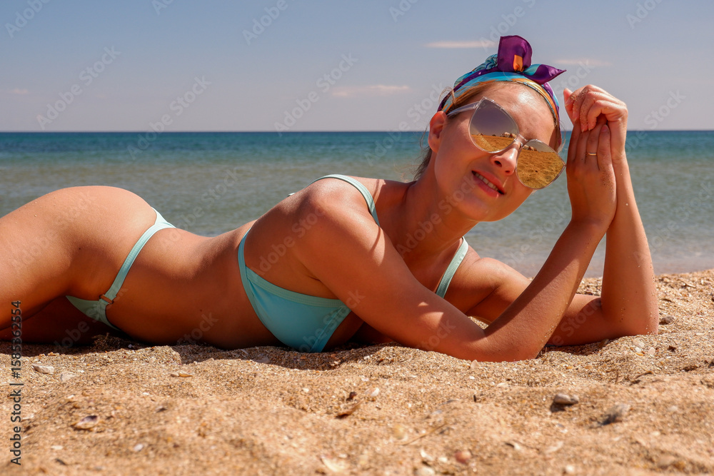 Young woman in blue bikini lying on sandy beach under sunlight, wearing sunglasses and colorful scarf. Relaxed beach vibe. Perfect for summer, vacation, travel, fashion, and lifestyle concepts.