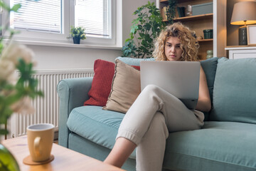 Concentrated millennial girl sit on couch working on laptop browsing internet at home during lazy weekend, focused young woman freelancer busy using computer surfing wireless web shopping online