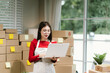 © Natthawadee - Young Asian woman in red shirt and apron doing online business, multitasking between smartphone and laptop, surrounded by packaging boxes in modern workspace.