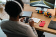© Studio Marmellata - An African American person wearing white headphones is engrossed in a video game. They hold a controller, playing a racing game displayed on a large monitor at their desk.