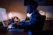 © Studio Marmellata - An African American man works intently on his laptop and large monitor in a dimly lit home office. He is focused on his task, illuminated by screen light.