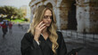 © Krakenimages.com - Young blonde woman in judge uniform stands outdoors near roman coliseum, using tablet and phone, embodying a modern legal professional setting.