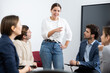 © JackF - Young female standing near near interactive board and sharing ideas during psychological trainings with colleagues sitting in circle on chairs