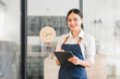 © kenchiro168 - Young woman wearing apron holding digital tablet smiling in cafe with open sign on glass door, bright atmosphere and friendly expression
