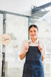 © kenchiro168 - Friendly young woman wearing apron standing inside cafe with open sign on glass door, smiling warmly inviting customers to come in, bright modern interior background, small business owner concept