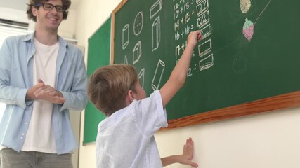 Poster - Happy caucasian boy writing answer at blackboard and giving high five while standing in front of classroom. Close up of smart elementary student volunteering to answer question. Education. Pedagogy.