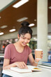 © Fahng - Female student sitting in cafeteria, reading book, studying for final exam at university.