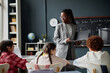 © Seventyfour - Black woman teacher holding file folder standing in front of classroom engaging with group of diverse children sitting at desks, listening attentively during science lesson about solar system