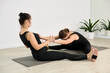 © TRAVELARIUM - Two young women practice partner yoga in studio. They sit on mats, stretching forward in synchronized pose. Light walls and plants create calming ambiance