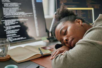 Poster - Young adult Black woman sleeping at desk with head resting on arms, surrounded by open notebook, pen, glass of water, and computer monitors displaying programming code