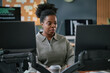 © Seventyfour - Young adult Black woman working at computer desk in modern office, focusing on dual monitors, coding software visible on screen in background
