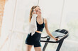© deagreez - Young woman cooling down after treadmill workout in bright gym, wearing stylish black activewear and holding towel