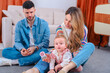 © alfons - Smiling parents sit on the living room carpet watching their curious baby daughter in pink overalls reach for a wooden cup, illustrating playful family bonding,early development and home joy together