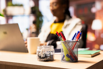 Poster - Focus on pens in desk organizer in front of remote worker typing on notebook keyboard. Close up of stationary items holder on home office desk next to teleworker using laptop