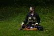 © arrideo - Happy smiling young man of Caucasian origin sitting on green grass background in a park. He is wearing a black kimono as a part of Chinese tea ceremony