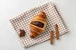© PutriNurAlleya - Croissant Resting on Checkered Napkin with Cinnamon Stick and Chocolate Square Overhead View