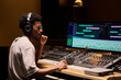 © pressmaster - Young adult Black man wearing headphones working at audio mixing console in professional recording studio, focusing on editing music tracks displayed on large computer monitors