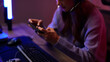 © STBSTD - Asian young adult woman streamer gamer playing mobile video game online using headset technology equipment in dark room glowing lights at night home setup