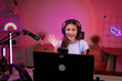© Pichsakul - Young Asian adult woman streamer content creator live streaming gaming online home room setup technology neon lights happy smile wave camera