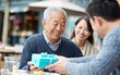 © Fathor - A young Asian man is handing out blue gift boxes to an elderly Asian father sitting at a table in an outdoor cafe. The father's daughter is standing behind him, smiling at her dad. This is an emotiona