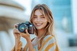 © k - Young woman smiling outdoors holding a camera up to her eye, ready to take a photo, with soft natural light and blurred urban background