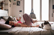 © Studio Marmellata - Side view of young tattooed female browsing smartphone and touching cat while resting on bed in light bedroom in morning at home