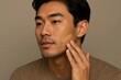 © Loginova - Young Asian man applying liquid foundation to his cheek, demonstrating men's skincare and grooming routine against a neutral background.