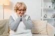 © Serhii - Mature woman sitting on sofa, touching inflammation lymph nodes, feeling discomfort and throat sore. Sick female with respiratory infection symptoms or chronic tonsillitis