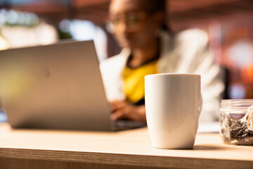 Poster - Focus on coffee mug on desk in front of remote worker typing on notebook keyboard. Close up of coffee cup on home office desk next to teleworker using laptop device to do job