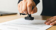 © synthetic - A person's hand holding a black rubber stamp over a document on a light brown wooden surface table