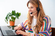 © Raquel - Smiling young woman in vibrant crochet sweater sits at a desk wearing a headset, typing on a laptop while engaged in a video call or online meeting, representing remote work and customer support.