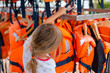 © kittyfly - little girl choosing life jacket for boating, orange life vests hanging, Safety equipment water sports, Summer activity preparation, water safety, adventure, and summer recreation, aquatic fun