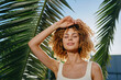 © SHOTPRIME STUDIO - smiling woman with curly hair posing outdoors near palm leaves under bright sunlight in summer, wearing a light tank top, natural beauty and relaxed mood