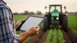 © nunoi - A person uses a tablet in a field, with a tractor in the background, showcasing modern agriculture technology.