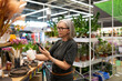 © Ivan Traimak - Stylish mature woman shopping for home goods at a modern retail store, selecting decor and plants in a vibrant environment