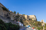 Fortified historical structure on a rocky hill under blue skies, with trees and modern road signs foregrounding the scene, showcasing architectural heritage and nature.