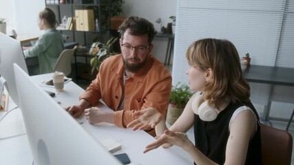 High angle shot of two male and female programmers sitting at wooden desk with two computers and office supplies while discussing new app glancing at monitor occasionally