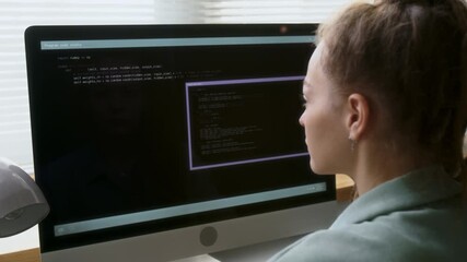 Over shoulder view of young woman sitting at desk and looking at monitor with codes