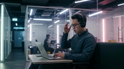 Poster - asian man wearing headphones working on laptop in modern office space. office worker typing on compu
