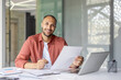 © Liubomir - A smiling man sits at a desk, holding documents with a laptop nearby. He appears to be reviewing paperwork.