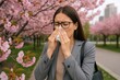 © Marica - Young professional woman experiencing springtime hay fever while working outdoors in blooming urban garden surrounded by flowering cherry trees