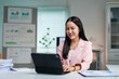 © amnaj - Asian businesswoman is smiling while working with a digital tablet at her desk in the office, with financial documents and charts in the background