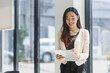 © amnaj - Young Asian businesswoman smiling and taking notes on a clipboard while standing next to a window in her office, she's wearing a white jacket and black trousers