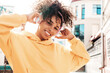 © halayalex - Beautiful woman with afro curls hairstyle.Smiling model in yellow hoodie.Sexy carefree female enjoying listening music in wireless headphones.Posing on street background at sunset
